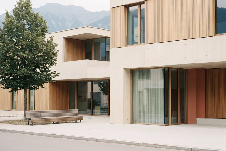 Façade claire en béton et bois d’un cabinet de santé de quartier en Suisse, avec un banc et un arbre.
